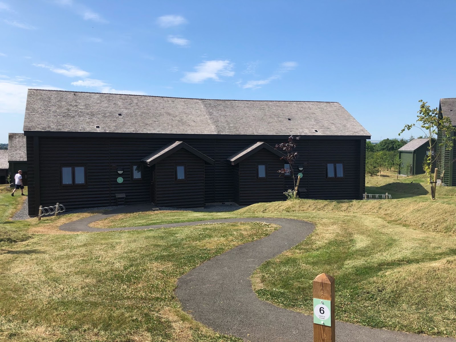 A dark wooden lodge-style building with a sloped shingle roof set on a grassy area with a curving paved path, under a bright sky.