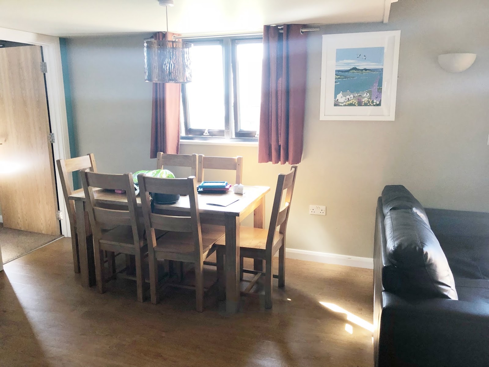 A dining room interior with a wooden table and six chairs, dark leather sofa, framed coastal artwork on wall, warm sunlight