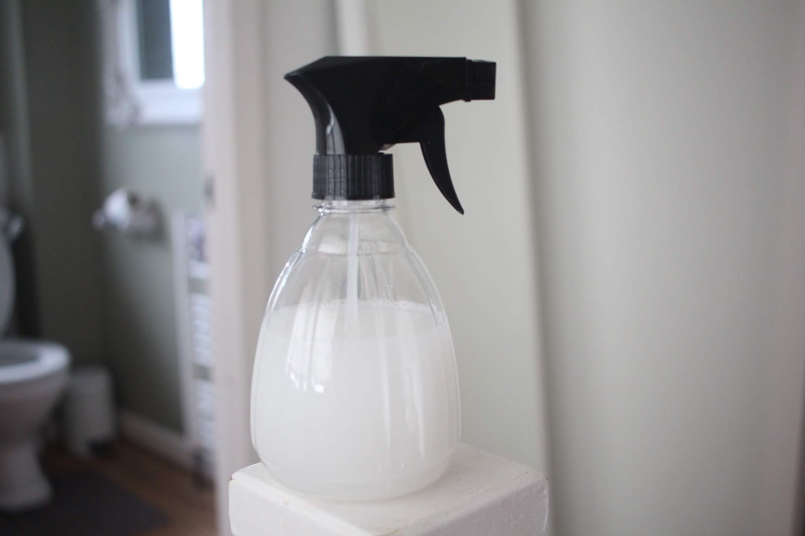 A clear spray bottle with a black trigger nozzle filled with white liquid, sitting on a white surface in soft bathroom lighting.